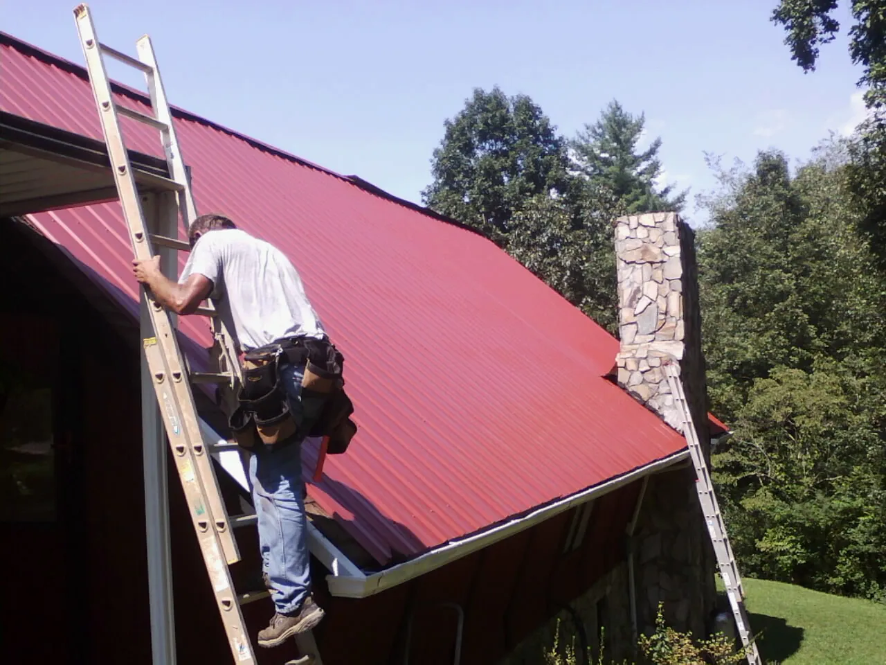 Roofer on a ladder beside a red metal roof and stone chimney — Soffit & Fascia Repair by Roofing Brothers in Mangum